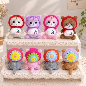 May include: Eight small crocheted cat plushies arranged on a two-tiered white stand. The top row features four cats wearing hoods with cherry embroidery. The bottom row features four cats wearing colorful, flower-shaped hoods with tails visible.