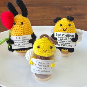 May include: Three handmade crochet bee figurines. One bee holds a red flower, another has white wings, and the third has a pair of wings. Each bee holds a sign with positive messages. The bees are yellow and black.