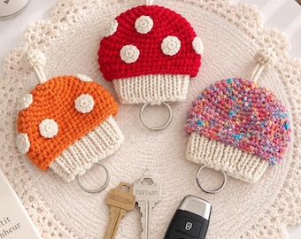 May include: Three crochet mushroom keychains with cream-colored stems and caps in red, orange, and multicolored yarn, each featuring white spots, a small top loop, and a metal ring. Two metal keys and a black car key fob are placed below them.