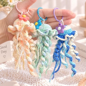 May include: Three crocheted jellyfish keychains in pastel peach, mint green, and blue and white. Each jellyfish features a round head, ruffled edges, and long, spiraling tentacles. A small, colorful flower charm is attached to each plastic ring.