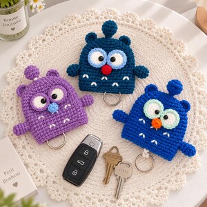 May include: Three crochet owl-shaped key holders in dark blue, purple, and bright blue. Each features large white eyes with black pupils, a small round nose, and a metal ring at the bottom. A black car key fob and two metal house keys are placed nearby.