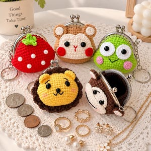 May include: Five crocheted coin purses with metal kiss-lock clasps and keychains. Designs include a red strawberry with white pearls, a tan bear, a green frog, a yellow lion, and a brown monkey. Coins, rings, earrings, and a necklace are nearby.