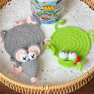 May include: Crocheted grey mouse and green frog coasters with white eyes and red tongues. A white mug with a colourful landscape design sits on a white doily inside a woven basket. Handmade home decor.