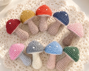 May include: A collection of ten small, handmade crochet mushrooms in various colors including red, yellow, dark blue, pink, gray, light blue, and green. Each mushroom features a textured cap with small white spots and a light-colored stem.