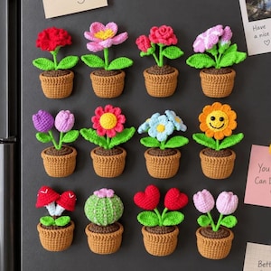 May include: A collection of twelve crocheted potted plant magnets arranged in three rows. Each features a brown textured pot with green leaves and various colorful flowers, cacti, or mushrooms, including one yellow sunflower with a smiling face.
