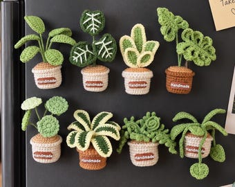 May include: Eight crochet plant magnets arranged in two rows. Each plant features green leaves in a small beige or brown pot with a brown tag labeled Handmade. The plants vary in leaf shape, including round, pointed, and textured foliage designs.