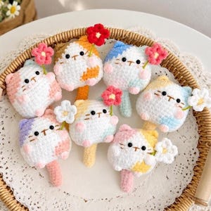 May include: Seven small, round, crocheted cat figures in various pastel colours, each featuring black bead eyes, a small embroidered mouth, and pink cheeks. Each cat holds a tiny crocheted flower in red, pink, or white and has a small tail.
