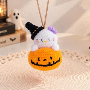 May include: A crocheted hanging ornament featuring a white character with pink cheeks and a purple bow, wearing a black witch hat with an orange band, sitting inside a bright orange pumpkin with a black stitched jack-o'-lantern face.