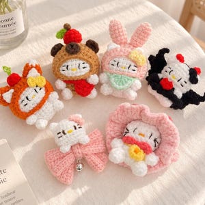 May include: Six small, handcrafted crochet figures featuring white cat-like faces with yellow noses and black eyes. Each figure wears a different colorful costume, including a red-topped brown bear, a pink bunny, a black bat, and various floral or bow accents.