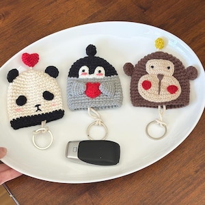 May include: Three handmade crochet keychains shaped like a panda, penguin, and monkey. The panda keychain is white and black with a red heart. The penguin is gray and black with a red heart. The monkey is brown with a beige face.