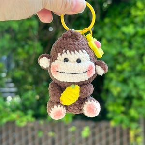 May include: A brown and cream crocheted monkey keychain featuring a yellow banana held in its hands, pink cheeks, and pink paw pads. The monkey is attached to a bright yellow plastic ring with a small yellow flower charm with a pink center.