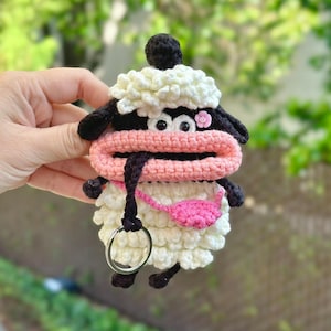 May include: A handmade crochet sheep keychain. The sheep has a white body, black ears and feet, and a pink mouth. A small pink flower is attached to the head. The keychain includes a silver metal ring.