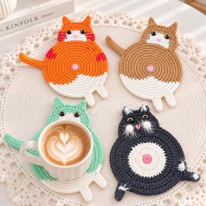 May include: Four round crochet cat coasters in orange, brown, mint green, and black. Each coaster features a white belly, a small pink center detail, pointed ears, and a tail. A white ceramic mug filled with coffee sits on the mint green coaster.