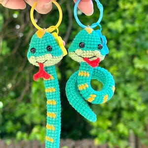 Peut inclure: Deux porte-clés serpent en crochet avec des corps en laine verte, des accents jaunes, des langues rouges et des yeux en perles noires. Un serpent pend droit tandis que l'autre est enroulé. Chacun est fixé à un anneau en plastique, jaune et bleu clair.