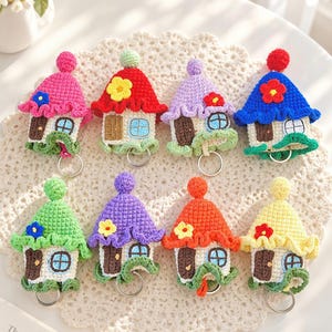May include: Eight crochet house keychains arranged in two rows. Each house features a pointed roof with a pom-pom, a brown door, a light blue window, and a small flower accent. The houses come in various colors including pink, red, purple, blue, green, orange, and yellow, each with a silver metal keyring attached at the bottom.