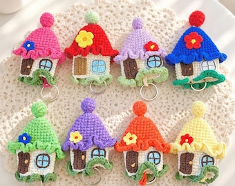 May include: Eight crochet house keychains arranged in two rows. Each house features a pointed roof with a pom-pom, a brown door, a light blue window, and a small flower accent. The houses come in various colors including pink, red, purple, blue, green, orange, and yellow, each with a silver metal keyring attached at the bottom.