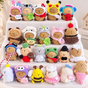 May include: A collection of twenty small, crocheted capybara figurines arranged on a tiered display. Each figure features a tan body and is dressed in various colorful, removable crocheted hoods and outfits, including animal, food, and character themes.