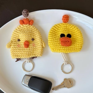 May include: Two handmade yellow crochet keychains shaped like chicks, each with orange beaks and accents. One has a small brown topknot and the other has black eyes. A car key and a house key are also in the image.