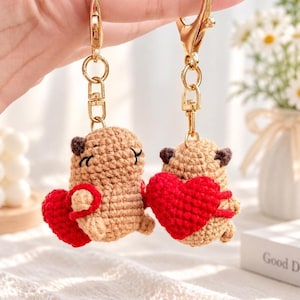 May include: Two tan crocheted animal keychains, each featuring a small brown-eared creature holding a bright red crocheted heart. The figures are attached to gold-toned metal lobster claw clasps and rings, showcasing detailed yarn stitching.