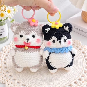 May include: Two crochet animal keychains featuring round bodies, black bead eyes, and pink cheeks. One is gray and white with a red collar and gold bell, the other is black and white with a blue ruffled collar. Both have plastic flower charms.
