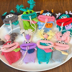 May include: A collection of colorful, handmade crochet monster-like creatures. Each has a unique color scheme, including green, blue, pink, and purple. They feature large mouths, googly eyes, and yarn accents, creating a whimsical and playful design.