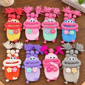 May include: Eight crocheted monster pouches in various colors including yellow, pink, mint, blue, orange, purple, and gray. Each pouch features two yarn pigtails, two plastic eyes, a wide mouth, and two small flower appliqués with pearl centers.