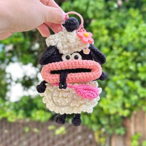 May include: A handmade crochet sheep keychain with a pink flower and a pink mouth. The sheep has black ears, eyes, and feet, and a white body. The keychain is held by a pink ring.