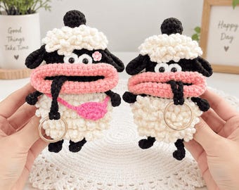 May include: Two handmade crochet sheep keychains featuring white woolly bodies, black heads, and prominent pink lips. One sheep wears a small pink crossbody bag and a tiny pink flower on its head. Both figures include a metal ring for attachment.