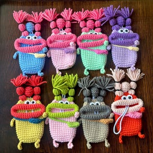 May include: A collection of colorful, handmade crochet monster-shaped pouches. Each pouch features a large, smiling mouth, googly eyes, and yarn hair. The pouches are in various colors, including blue, pink, green, yellow, and orange.