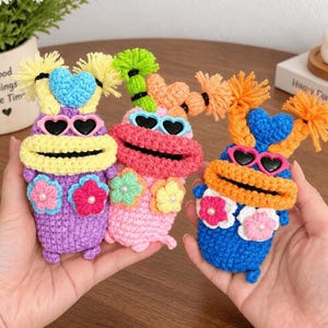 May include: Three small, hand-crocheted plush figures in purple, pink, and blue. Each features yarn hair, heart-shaped sunglasses, a wide mouth, and two crocheted flowers with pearl centers on the chest. The figures have textured yarn bodies.