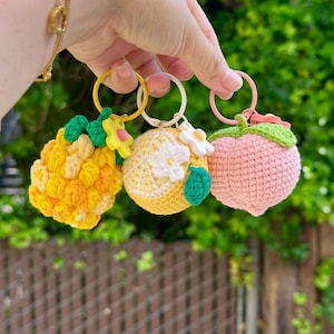 May include: Three crochet keychains: a yellow pineapple, a yellow lemon with a white flower, and a pink peach. Each keychain has a metal ring. The pineapple and lemon have green leaves. The peach has a green leaf and pink accents.
