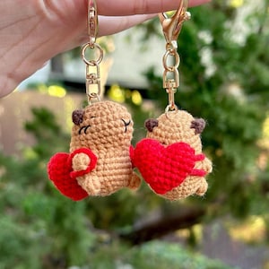 May include: Two crochet keychains featuring small, brown bear-like figures each holding a red heart. The keychains have gold-colored clasps. The bears have small, dark eyes and are made of a textured, tan yarn. The hearts are a vibrant red.