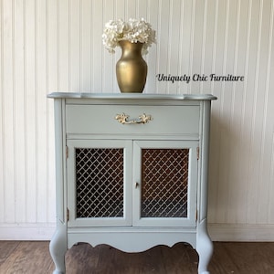 May include: A light blue painted cabinet with a drawer and two doors with wire mesh inserts. The cabinet has ornate gold hardware and curved legs. A gold vase with white flowers sits on top. The text "Uniquely Chic Furniture" is visible.
