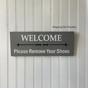 May include: Gray wooden sign with white text that reads "WELCOME" and "Please Remove Your Shoes". The sign has a black arrow pointing to the right.