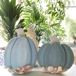 May include: Two blue painted wooden pumpkins decorated with seashells. The pumpkins are sitting on a white surface in front of a window.