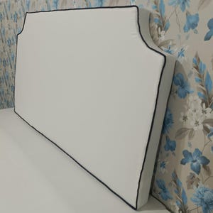 May include: A white headboard with a black trim, featuring a curved top edge. The headboard is positioned against a floral patterned wallpaper with blue and gray flowers.