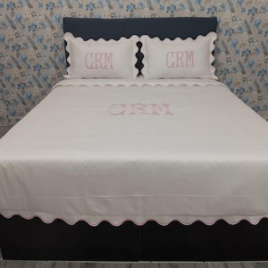 May include: White bedding set with pink scalloped trim and embroidered initials. The set includes a fitted sheet, a flat sheet, and two pillowcases. The initials "CRM" are embroidered on the pillowcases and the flat sheet. The bed is against a patterned wallpaper.