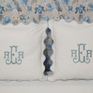 Puede incluir: Dos cojines cuadrados blancos con un borde festoneado y un monograma bordado en azul claro. Los cojines se exhiben sobre una superficie blanca contra un fondo con estampado floral con flores azules.