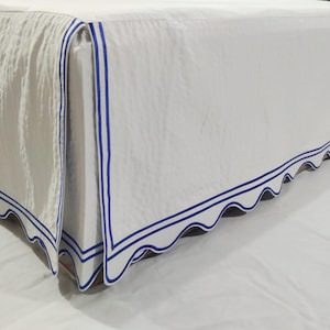 Może przedstawiać: Biała spódnica do łóżka z falistym brzegiem i podwójną granatową obwódką. Tkanina ma subtelną fakturę, a design dodaje łóżku klasycznego charakteru.