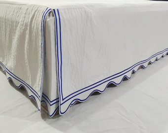 May include: A white bed skirt with a scalloped edge and a double navy blue border. The fabric has a subtle texture, and the design adds a classic touch to the bed.