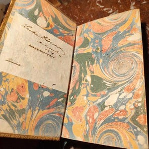 May include: Open antique book with marbled pages in shades of orange, blue, and green. The left page has handwritten text and a label. The book's cover is visible, showing a gold-colored trim. The book is open, revealing the intricate patterns.