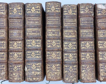 May include: A collection of antique books with brown leather covers. The spines feature gold lettering and decorative floral patterns. The books are labeled "Histoire Romaine" with volume numbers.