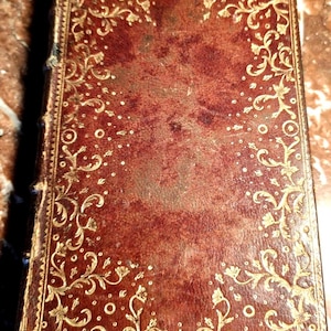 May include: An antique book featuring a reddish-brown leather cover, showing signs of wear. The cover is decorated with gold floral and foliate patterns. The spine of the book shows signs of age, adding to its vintage aesthetic.