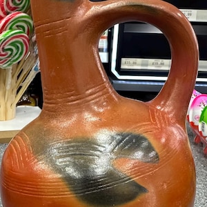 May include: A terracotta-colored clay pitcher with a rounded body, a tall neck, and a single arched handle. The surface features dark, smoky black markings and horizontal incised lines around the neck and body, resting on a woven base.