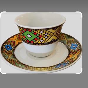 May include: White ceramic teacup and saucer set. The cup and saucer feature a colorful geometric pattern with red, green, yellow, and blue accents. The design is repeated on the cup and saucer.