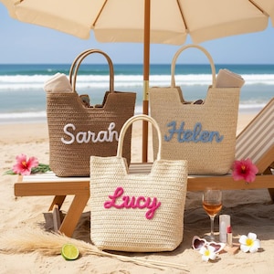 Op de afbeelding: Drie geweven draagtassen met handvatten, elk gepersonaliseerd met een naam: "Sarah" in wit, "Helen" in lichtblauw en "Lucy" in roze. De tassen staan op een strand met een parasol en een ligstoel.