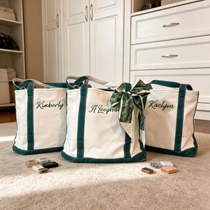 K&ouml;nnte beinhalten: Drei wei&szlig;e Canvas-Tragetaschen mit gr&uuml;nen Verzierungen und Griffen. Jede Tasche hat einen in Schwarz gestickten Namen: "Kimberly", "A Leeghan" und "Kaelym". Die mittlere Tasche hat einen gr&uuml;n gemusterten Schal am Griff. Kleine Schachteln befinden sich vor den Taschen.