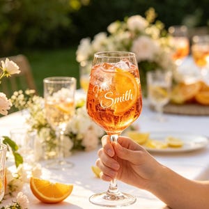 Op de afbeelding: Een helder wijnglas gevuld met een oranje cocktail, ijsblokjes en een verse schijf sinaasappel. Het glas is voorzien van de witte gegraveerde tekst "Mrs. Smith" op de zijkant. Het glas heeft een lange, slanke steel en een ronde voet.