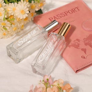 May include: Two clear glass perfume bottles with silver and gold caps, one labelled "Sophia" and the other "Charlotte The next chapter." A pink passport holder and flowers are in the background.