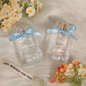 May include: Two clear glass perfume bottles with light blue satin ribbons tied around the necks. Each bottle features white floral line art, custom text including Sophia, Bridesmaid, Maid of the Honour, and the date 18.10.2025 printed on the front.